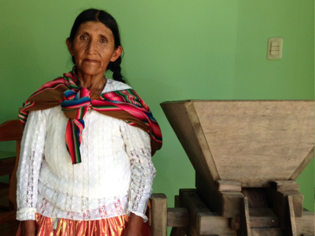 伝統的な衣装をまとったレアンドラさんが木製の脱穀機の隣に立つ